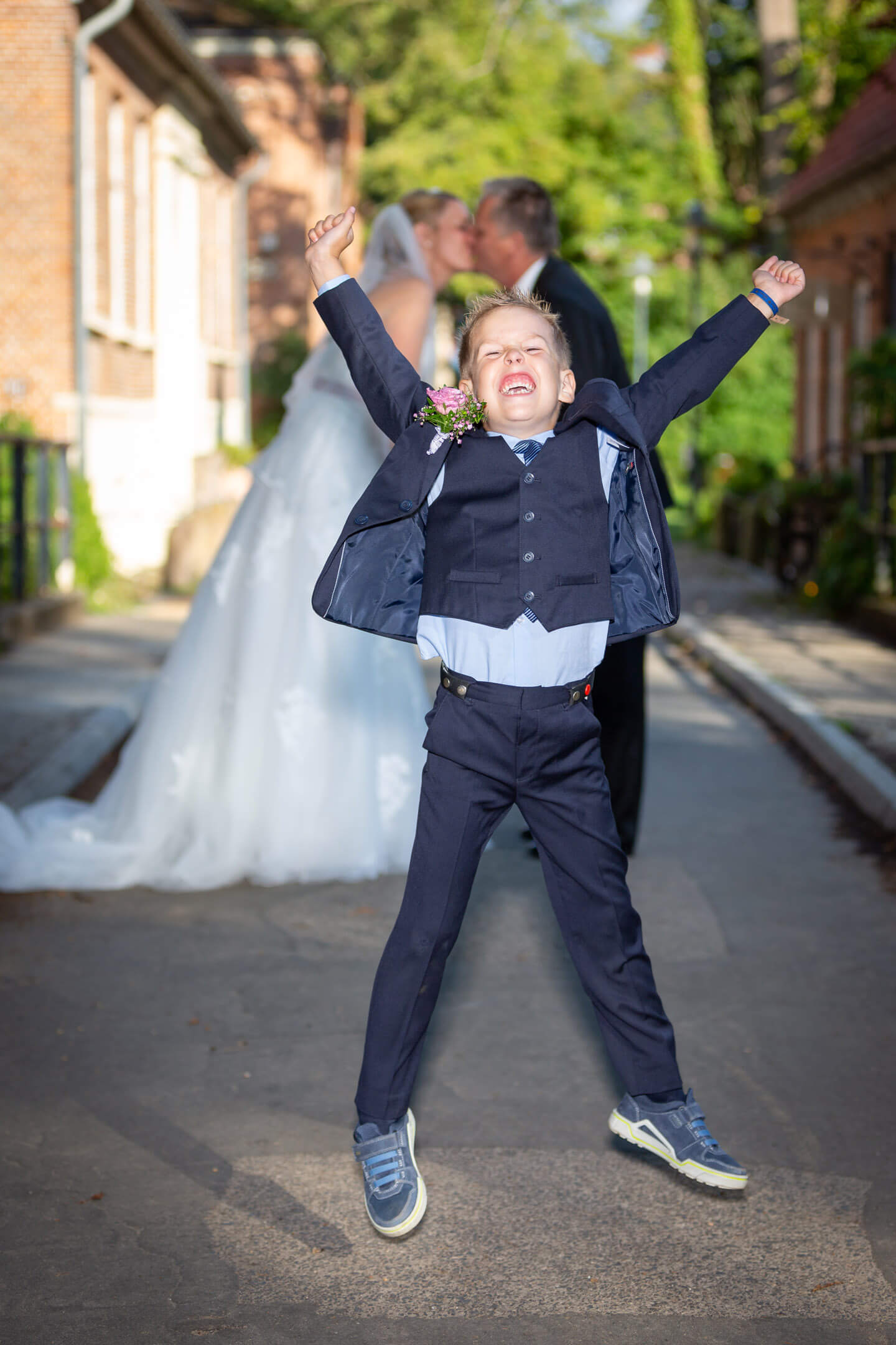Kind springt vor Freude in die Luft, im Hintergrund küssen sich die frischverheirateten Eltern