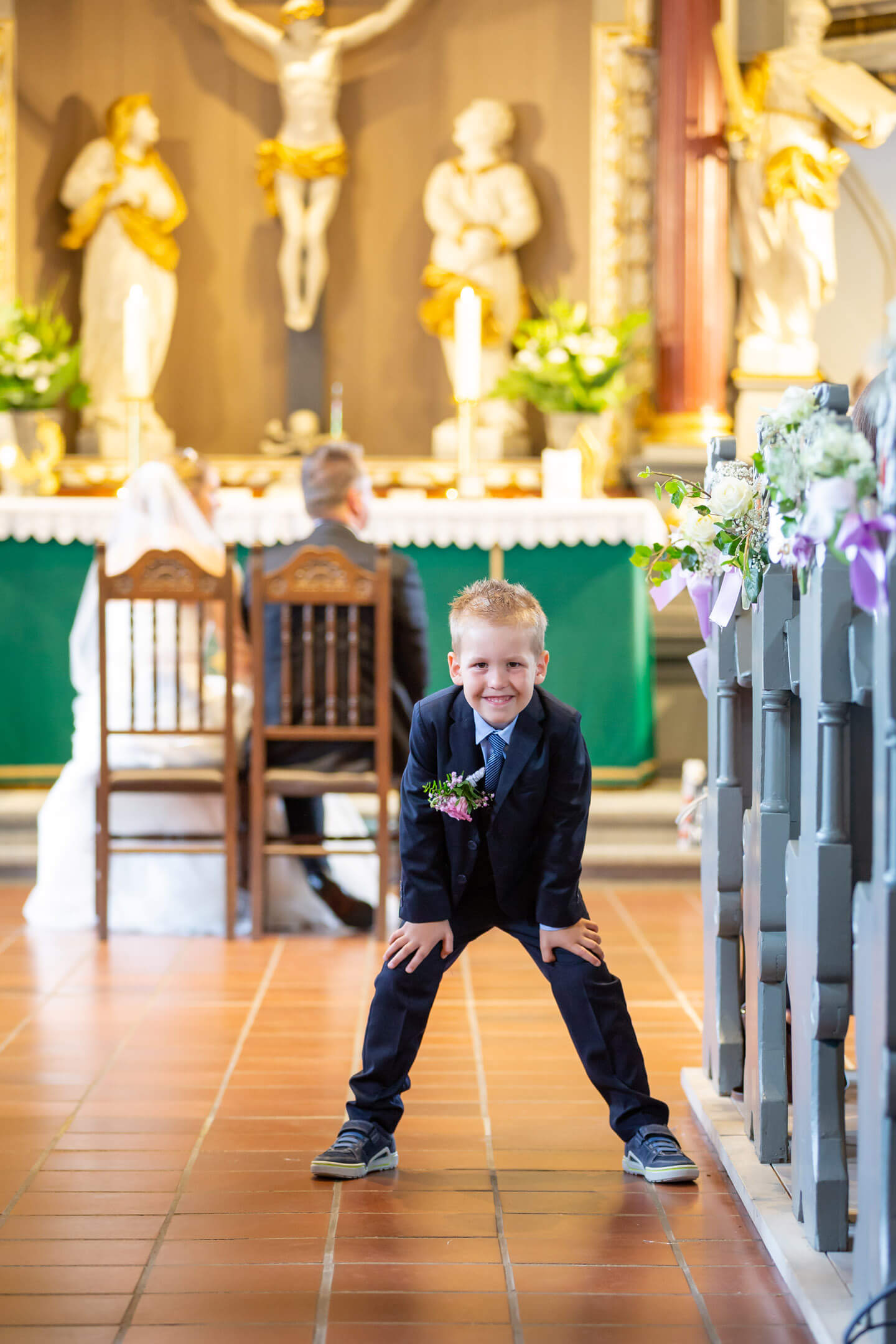 Kind guckt verschmitzt vom Kirchengang in die Kamera. Im Hintergrund sitzt das Brautpaar am Altar