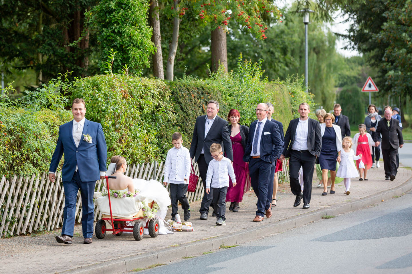 Bräutigam zieht die Braut im Bollerwagen zur Location. Hinter ihm die Hochzeitsgesellschaft