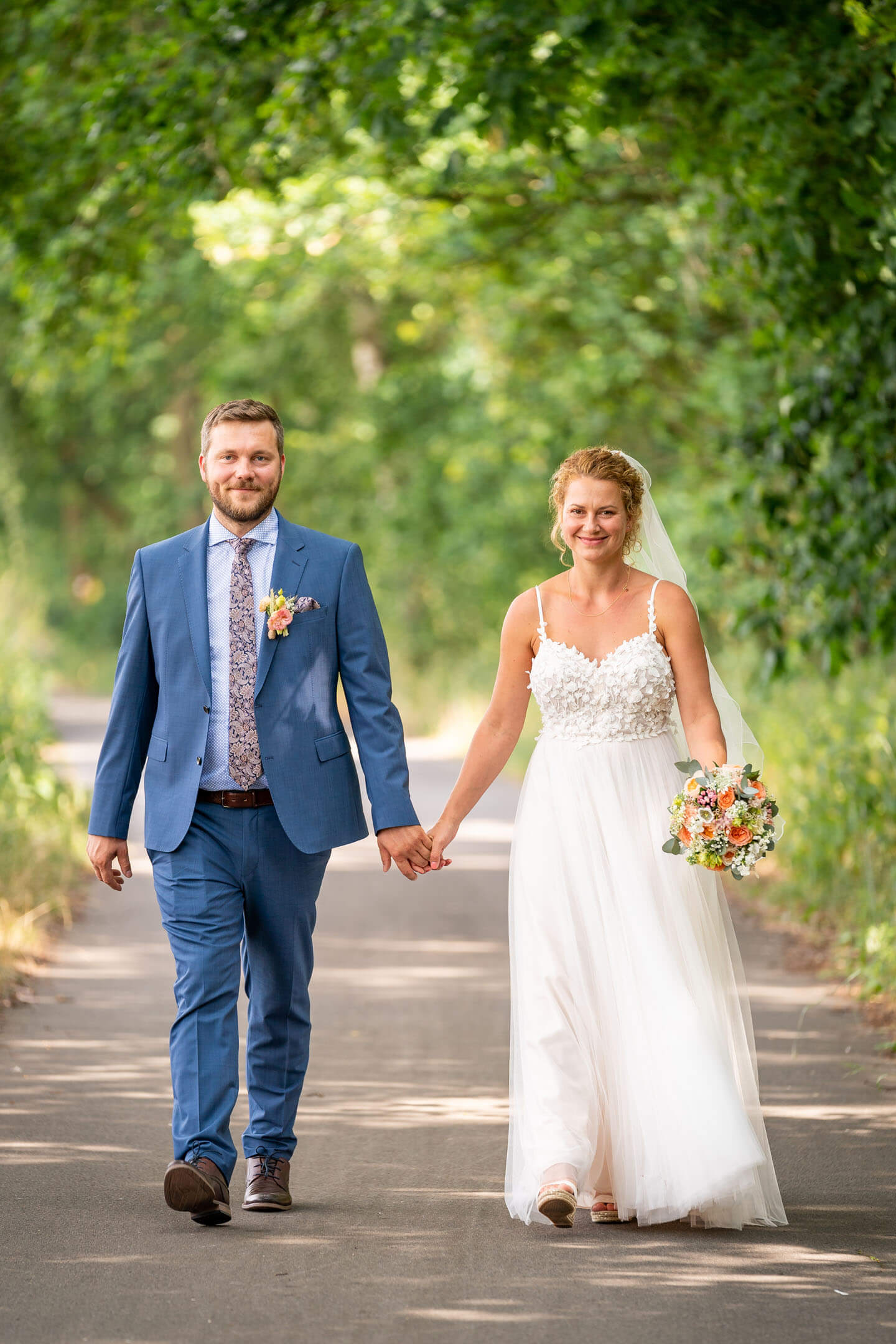 Hochzeitspaar geht Hand in Hand eine Allee entlang