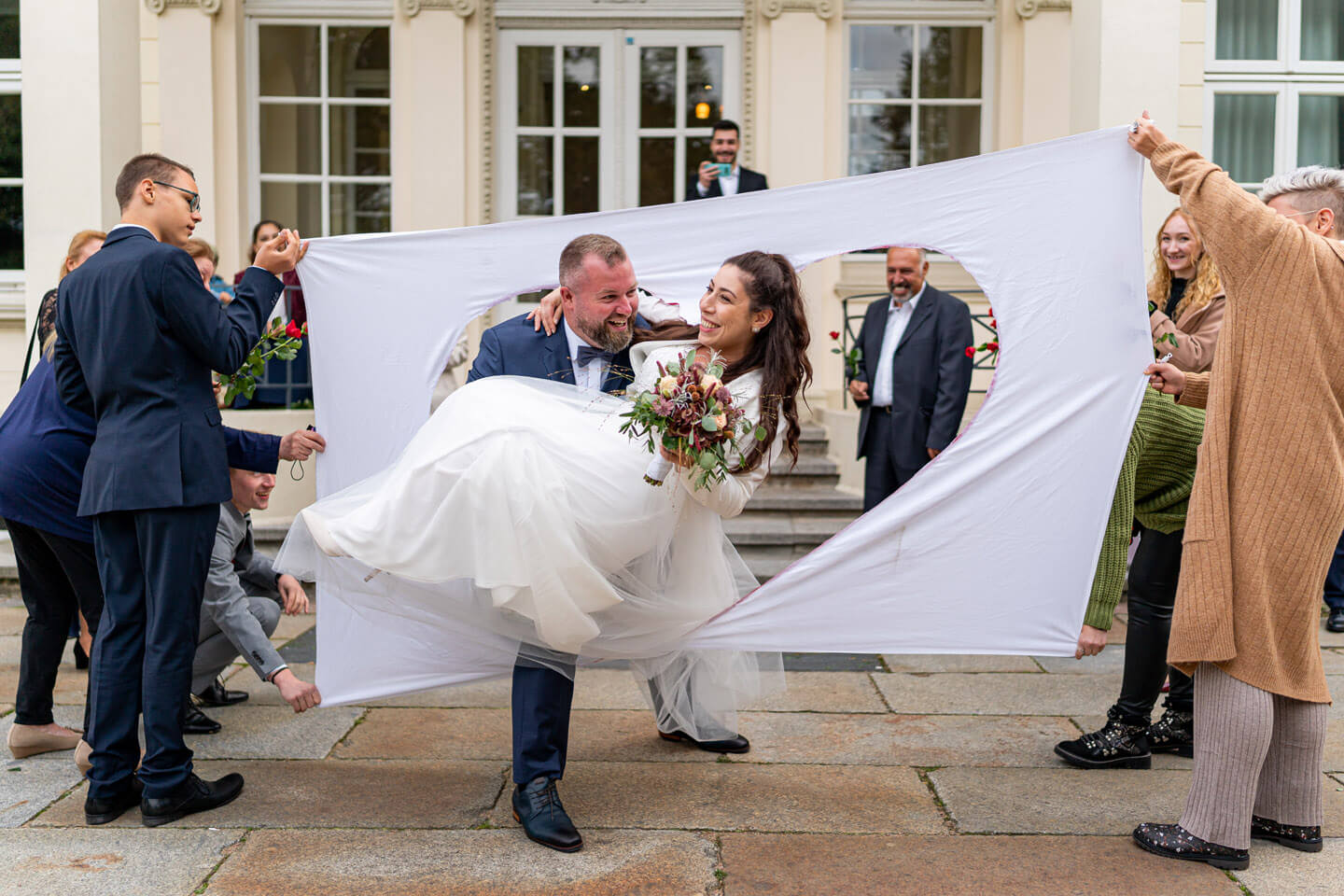 Bräutigam trägt Braut durchs ausgeschnittene Herz des Bettlakens.